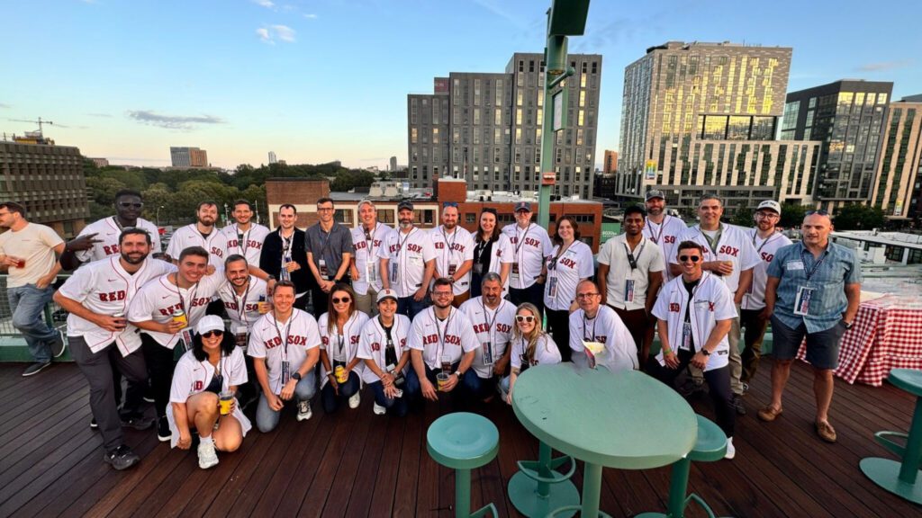A group of Braincube Go Beyond Summit 2025 attendees wearing Red Sox jerseys pose together on a rooftop deck at Fenway Park in Boston. The skyline and evening sky are visible in the background, capturing the relaxed, team-oriented atmosphere of the summer event.