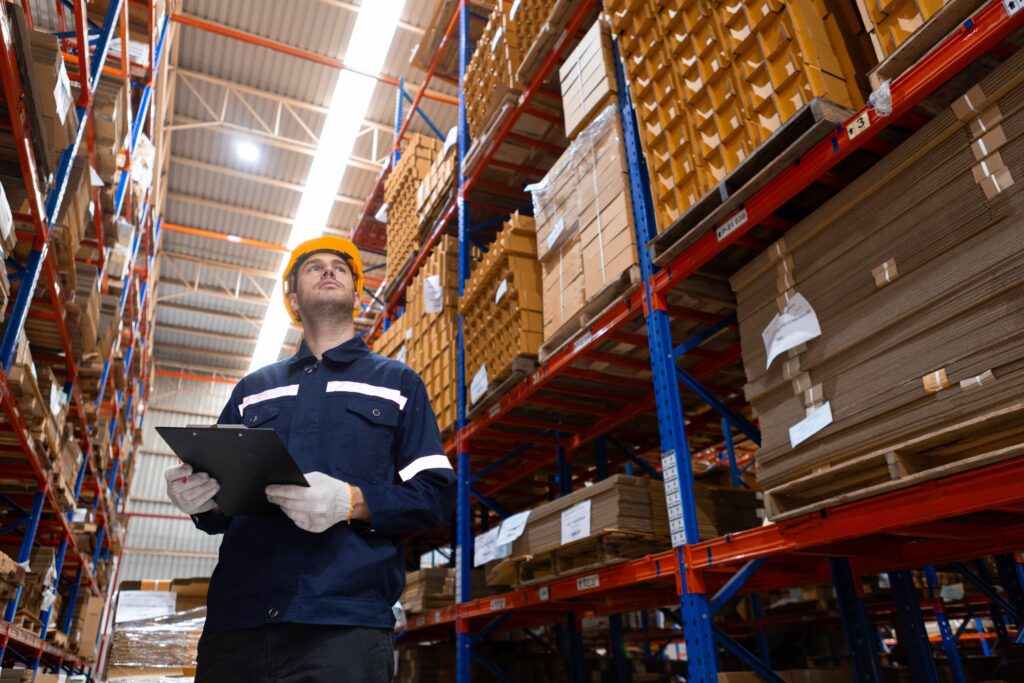 A factory worker wearing a hard hat and safety gloves stands in a warehouse aisle, holding a clipboard and inspecting tall shelves stacked with boxes and materials.